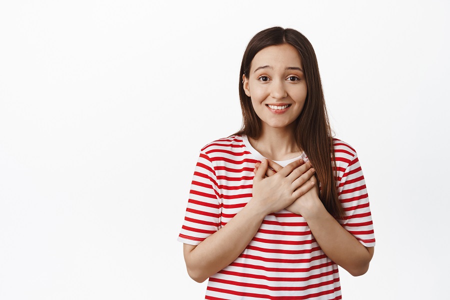 Young smiling girl looks thankful, holds hands on heart and thanking you, feel grateful, express gratitude and joy, standing against white background